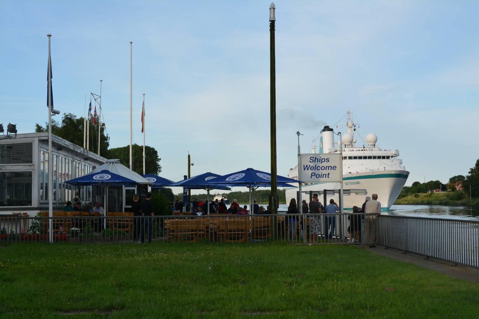 Rasenfläche mit Fahnenmasten vor der Terrasse, dahinter ein Schiff auf dem Nord-Ostsee-Kanal