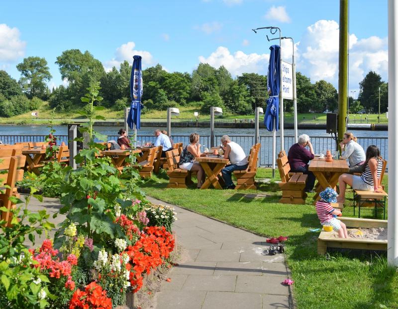 Biergarten mit Holzbänken und Blumenbeet am Kanal