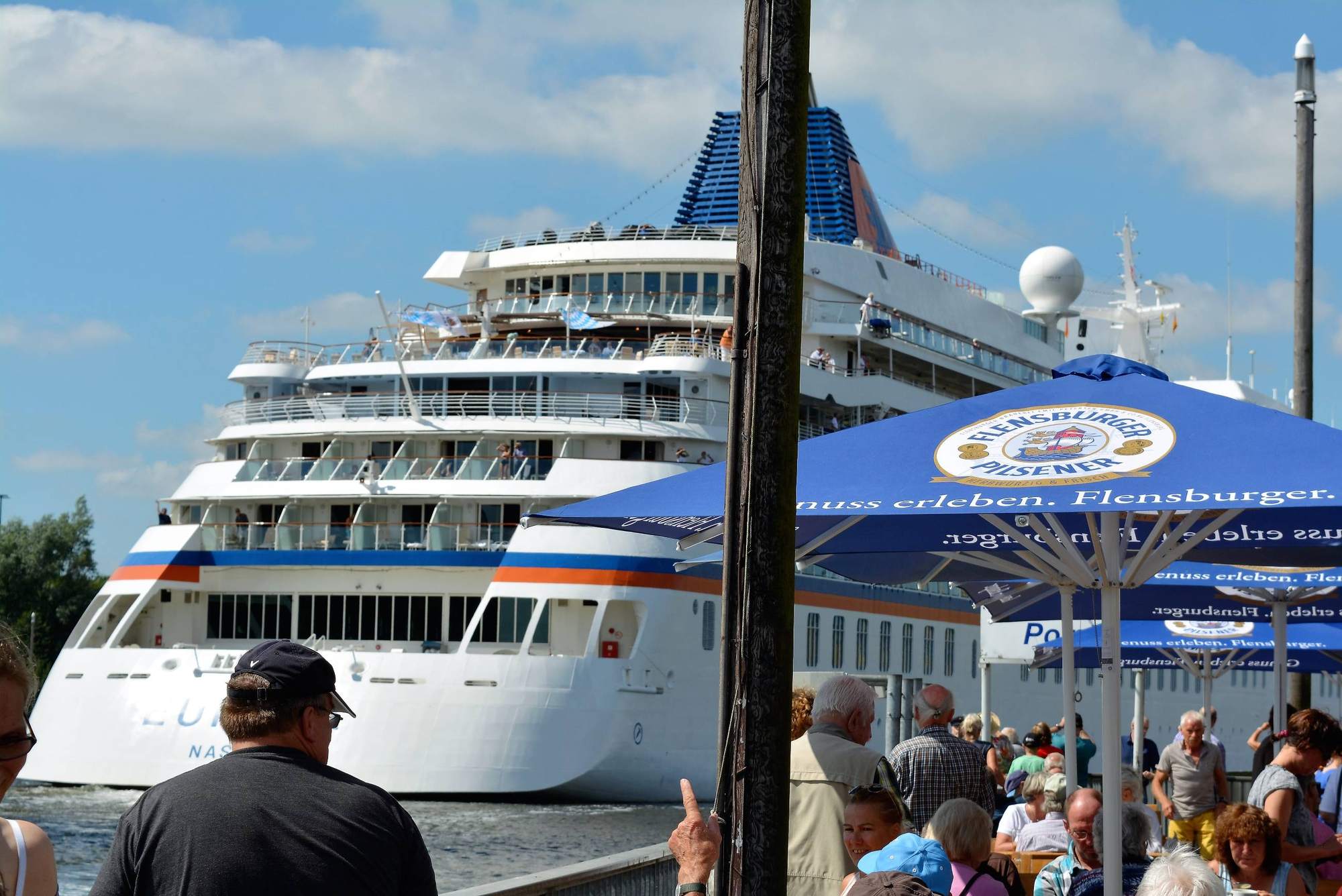 Gäste beobachten von der Terrasse aus ein weißes Kreuzfahrtschiff, das hinter den Sonnenschirmen vorbeizieht