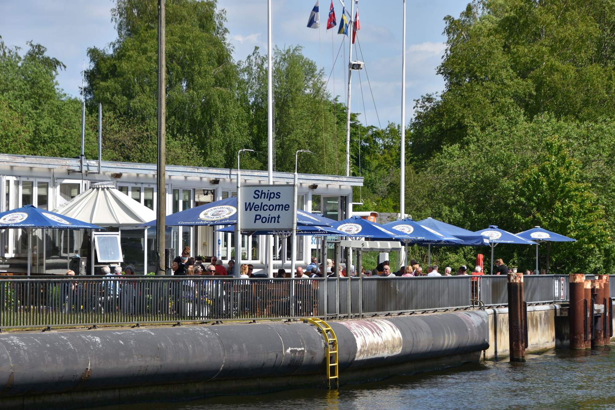 Terrasse der Brückenterrassen am Ufer des Nord-Ostsee-Kanals mit blauen Sonnenschirmen und dem Gebäude „Ships Welcome Point“